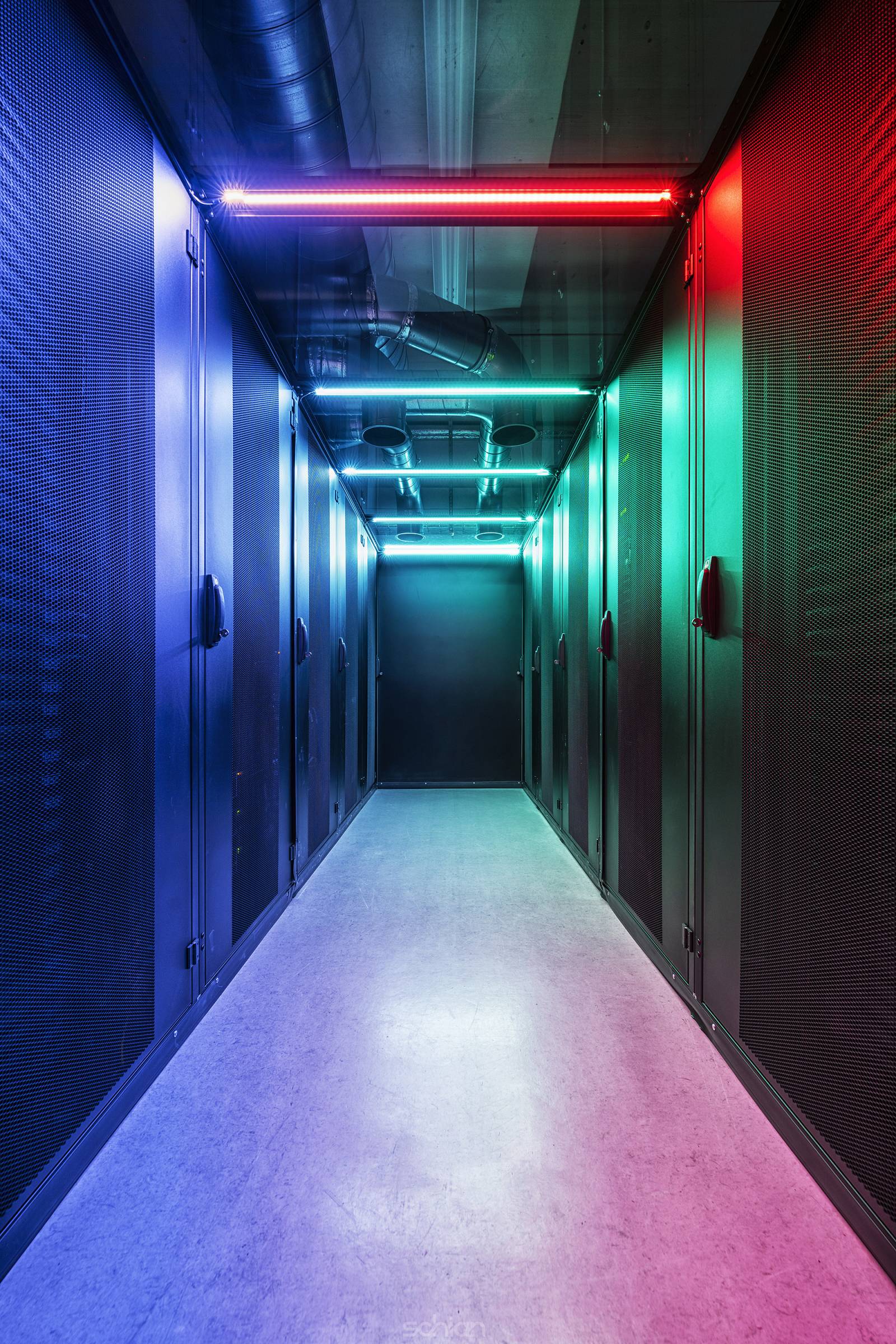 Cold aisle of an IT server room with perforated grille doors illuminated in color and large cooling pipes on the ceiling
